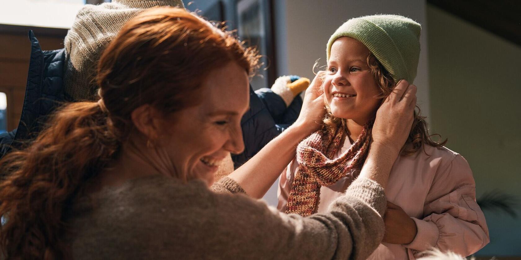 Eine Mutter zieht einem Mädchen eine Mütze an, beide lachen